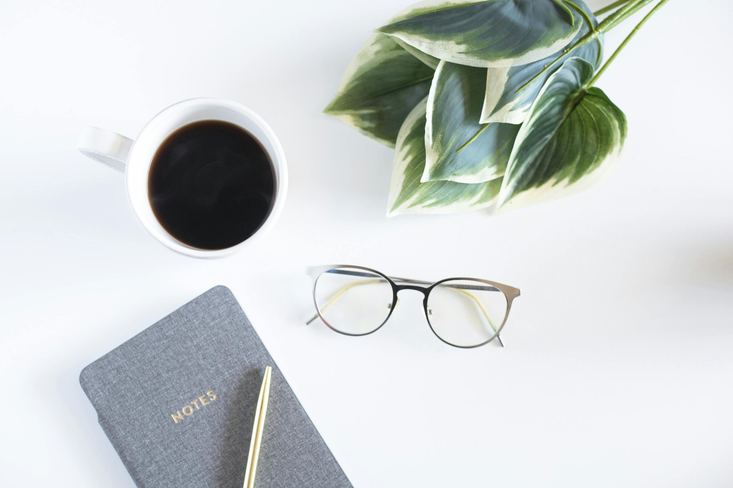 pexels-photo-5423285-5423285 A minimalist workspace featuring a coffee cup, notebook, and eyeglasses on a white desk.